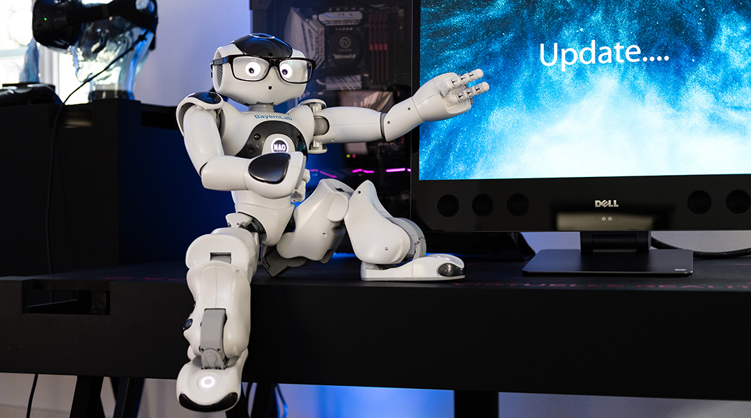 Ein humanoider Roboter sitzt auf einem Tisch vor einem Monitor. Der Roboter trägt eine schwarze Brille und zeigt mit ausgestrecktem Arm auf den Bildschirm, auf dem das Wort „Update…“ zu lesen ist. Im Hintergrund sind technische Geräte und Kabel zu sehen.